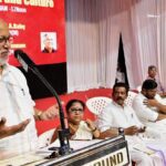 Indian left leaders speak at podium in front of red background at seminar