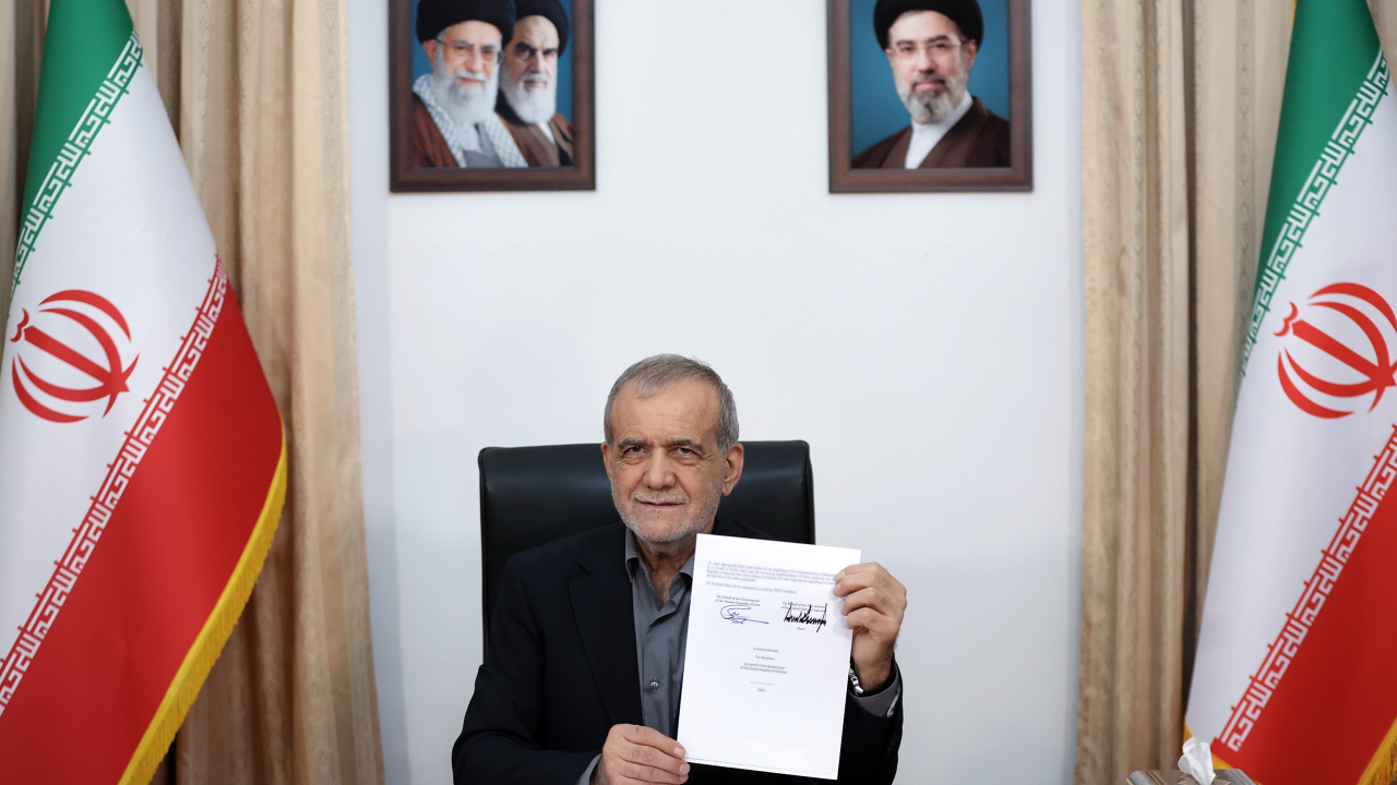 Iranian President Masoud Pezeshkian displays a document at a desk flanked by Iranian flags