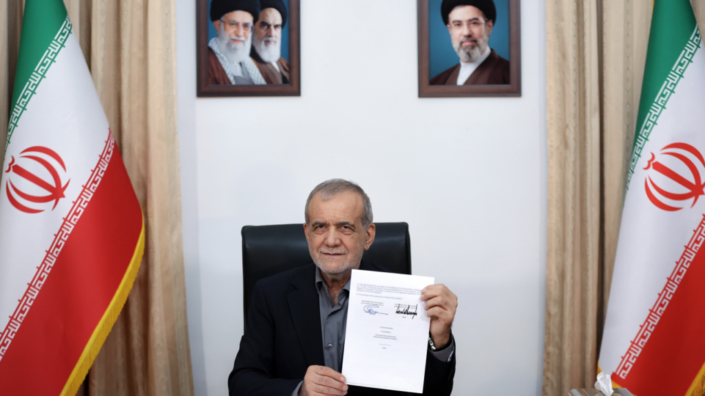 Iranian President Masoud Pezeshkian displays a document at a desk flanked by Iranian flags