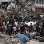 Child looks out at a crowd moving bodies in bags amidst rubble